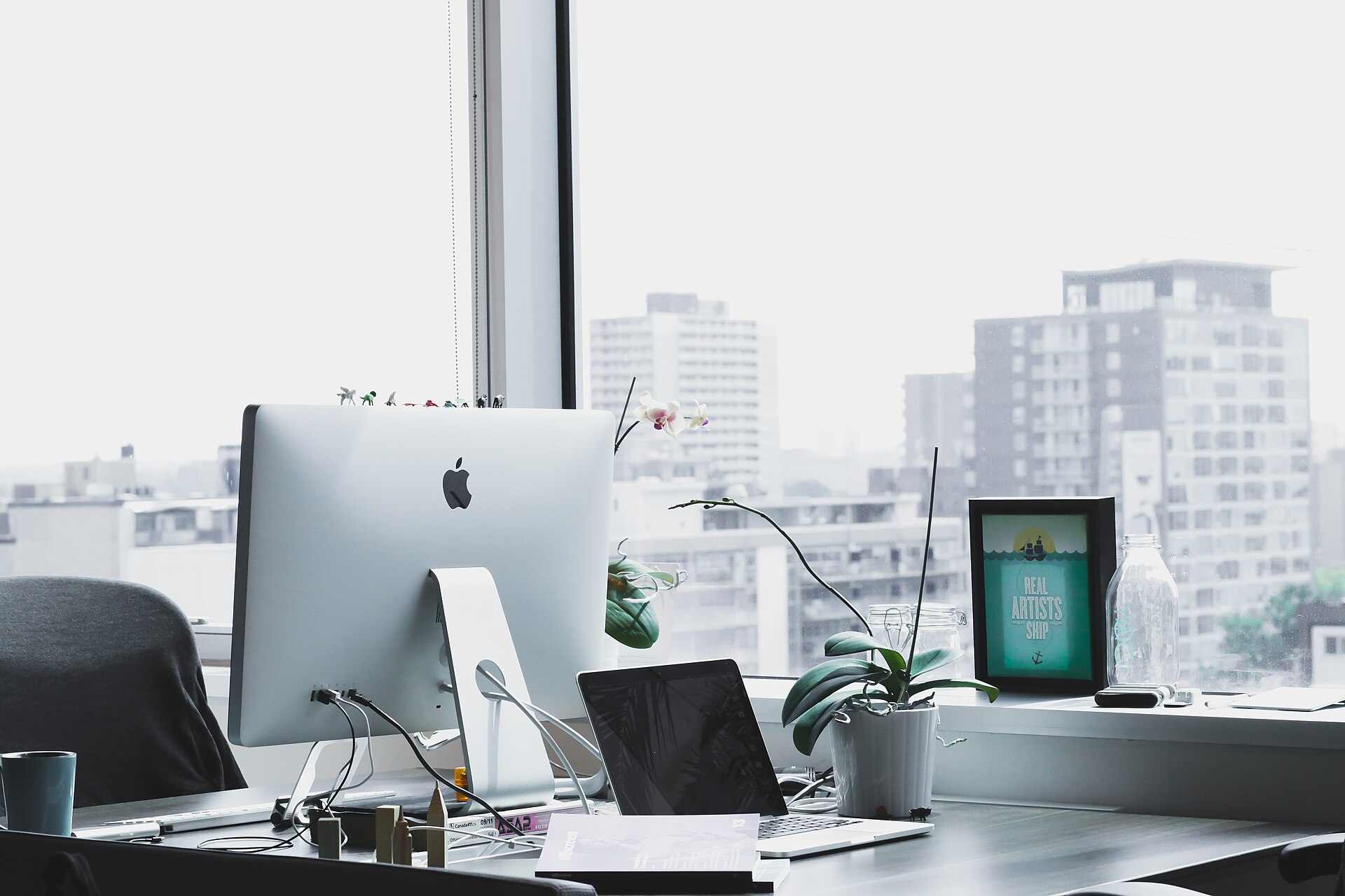 A bright workspace with a desktop monitor, laptop and a small plant by a window